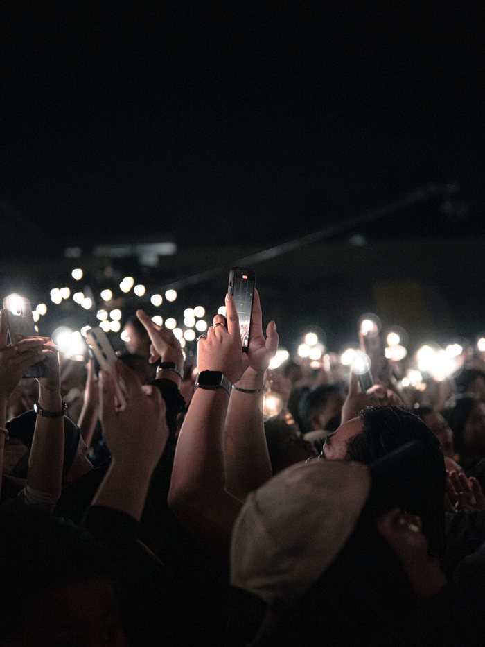 A crowd in the dark with phones held up, a field of lit screens carrying the same moment